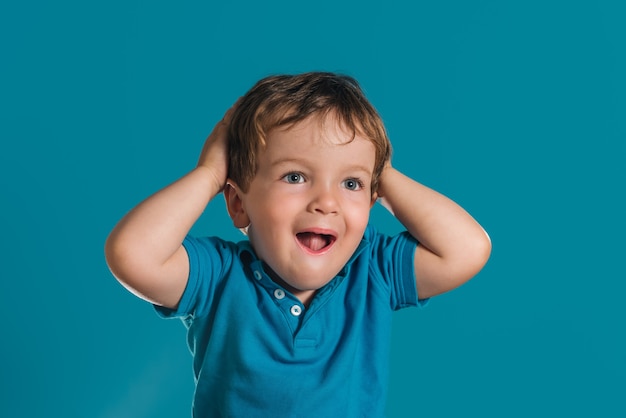 Close de um garotinho de camisa azul, colocando as mãos na cabeça, surpreso com um fundo azul