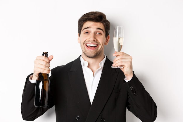 Foto close de um homem bonito de terno, segurando uma garrafa e uma taça de champanhe, comemorando o feriado, em pé contra um fundo branco