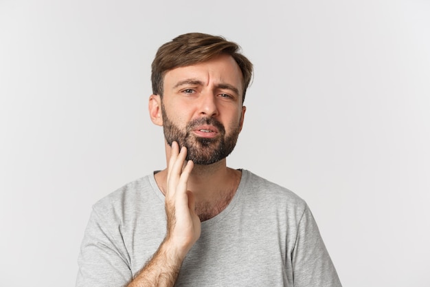 Close de um homem confuso tocando a barba e olhando para si mesmo, precisa se barbear, em pé sobre o branco.