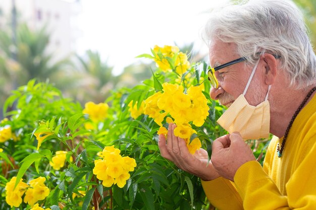 Close de um homem sênior sorridente, tirando a máscara cirúrgica ao ar livre devido a um coronavírus, cheirando uma flor amarela