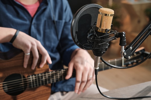 Close de um homem sentado em frente ao microfone tocando violão no estúdio