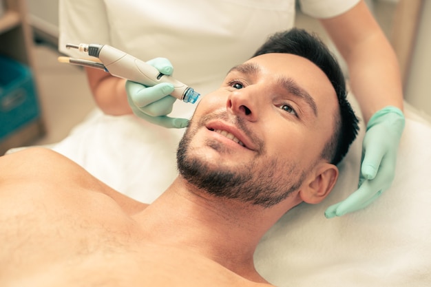 Close de um jovem barbudo parecendo feliz com uma ferramenta moderna perto de seu rosto para nutrir a pele