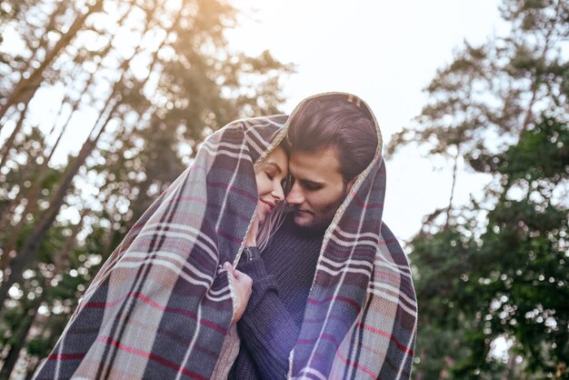 Close de um jovem casal feliz rindo debaixo do cobertor em um dia frio com uma floresta de outono ao fundo