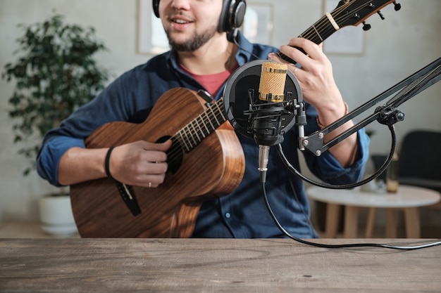 Close de um músico sentado em frente ao microfone tocando violão no estúdio de gravação
