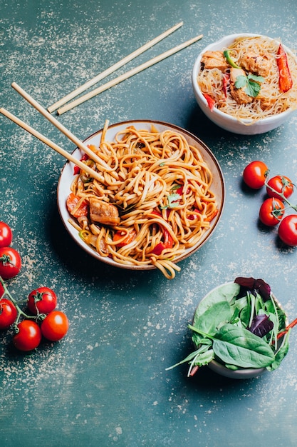 Close de um prato de macarrão chinês com carne e vegetais em pratos e pauzinhos chineses em uma superfície verde