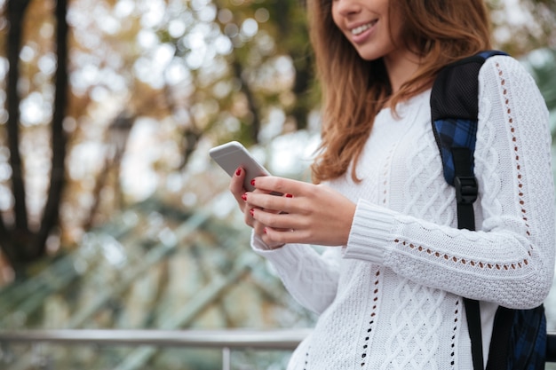 Close de uma bela jovem feliz com uma mochila usando o telefone celular no parque