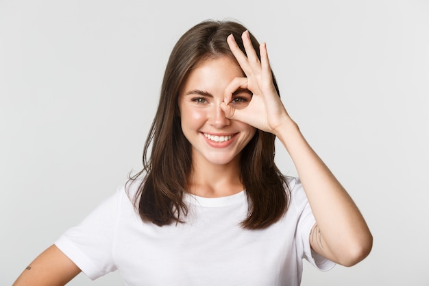 Close de uma garota atraente e confiante sorrindo satisfeita e mostrando um gesto de aprovação sobre o olho