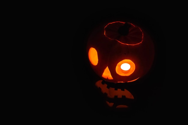 Foto close de uma jack o' lantern acesa no escuro e refletida em uma superfície preta à sua frente