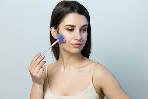 Foto close de uma jovem com um top claro sobre um fundo branco fazendo uma maquiagem facial