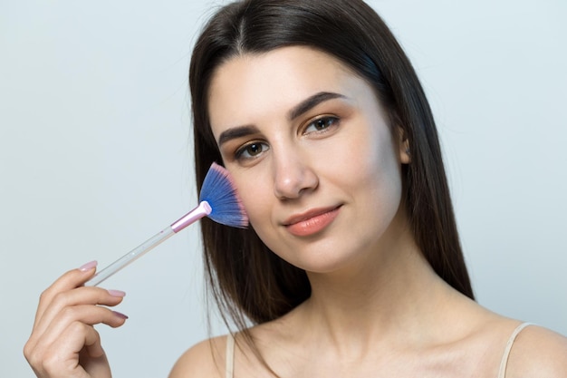 Close de uma jovem em um top leve sobre um fundo branco, fazendo uma maquiagem facial Uma mulher bonita segura um pincel cosmético perto do rosto e sorri