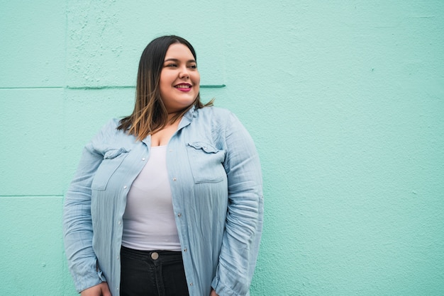 Close de uma jovem mulher plus size sorrindo em pé contra uma parede azul claro ao ar livre