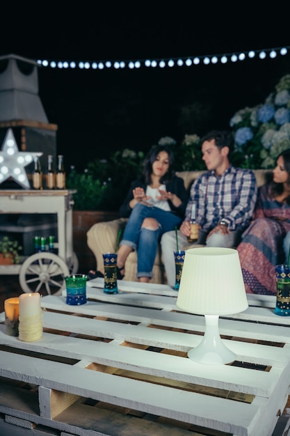Close de uma lâmpada branca sobre uma mesa de paletes em uma festa ao ar livre com pessoas conversando ao fundo