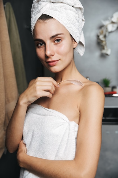 Foto close de uma linda jovem com uma toalha enrolada na cabeça e tocando sua pele com uma pena no banheiro