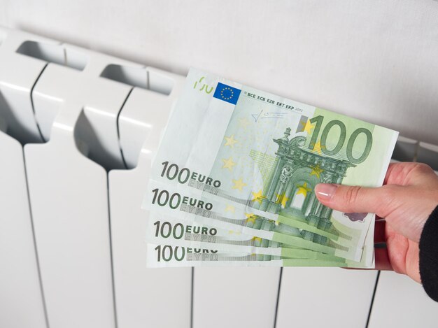 Close de uma mão segurando notas de cem euros No fundo, a bateria de aquecimento central é branca O conceito de pagamentos elevados na Europa aumenta nas contas de serviços públicos