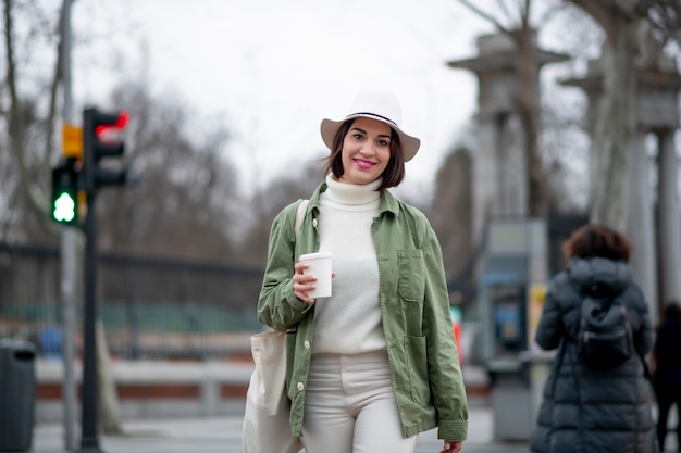 Foto close de uma mulher com chapéu branco tomando café enquanto caminha pela rua