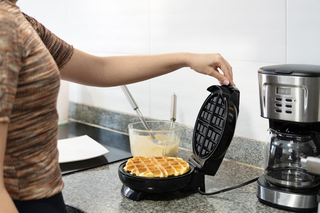 Close de uma mulher cujo rosto não é visível preparando waffles dourados na máquina de waffle ao lado da cafeteira