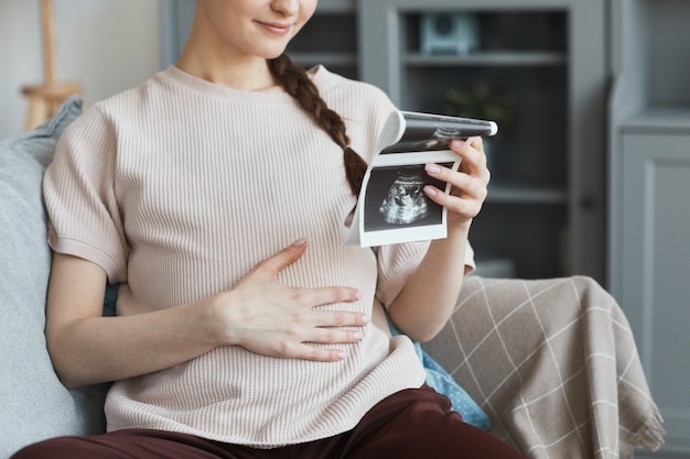 Close de uma mulher grávida sentada no sofá na sala e olhando para a imagem do raio-x do bebê