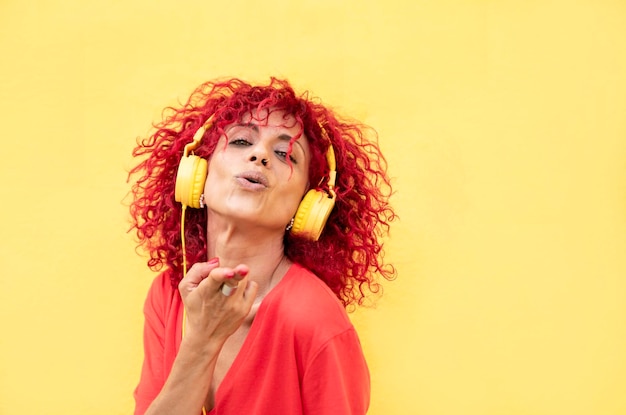 Close de uma mulher latina sorridente com cabelo afro vermelho ouvindo música com fones de ouvido amarelos em um fundo amarelo olha para a câmera e manda um beijo com as mãos