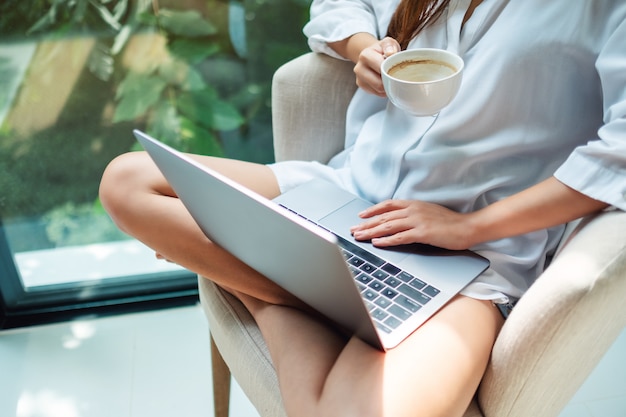 Close de uma mulher que trabalha no computador portátil enquanto bebe café e sentado em uma poltrona branca em casa com a natureza verde