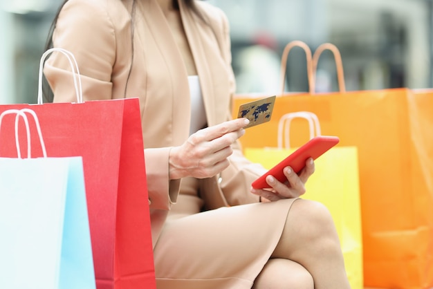 Close de uma mulher segurando um celular e um cartão de crédito perto de sacolas de compras multicoloridas