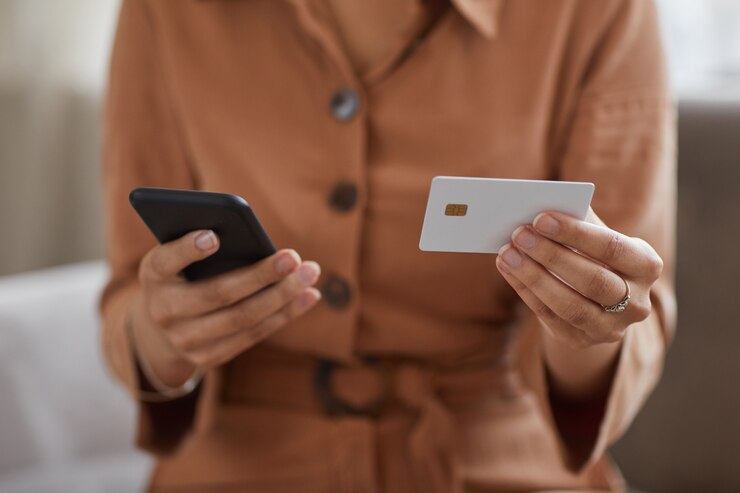 Close de uma mulher segurando um telefone celular e um cartão de crédito nas mãos, ela pagando contas online