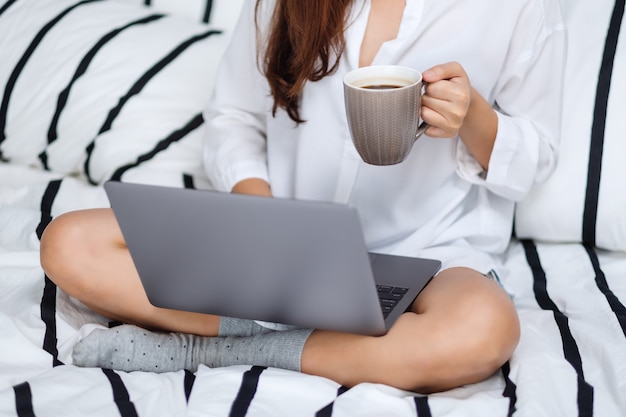 Close de uma mulher usando e trabalhando no computador portátil, bebendo café enquanto está sentado em uma cama branca e aconchegante em casa