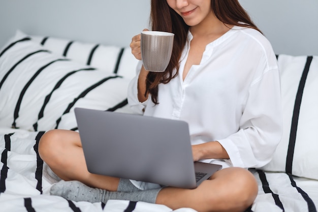 Close de uma mulher usando e trabalhando no computador portátil, bebendo café enquanto está sentado em uma cama branca e aconchegante em casa