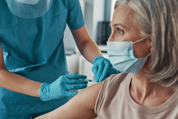 Close de uma paramédica com roupa de trabalho de proteção fazendo a vacinação contra o coronavírus em uma mulher madura enquanto trabalhava no hospital