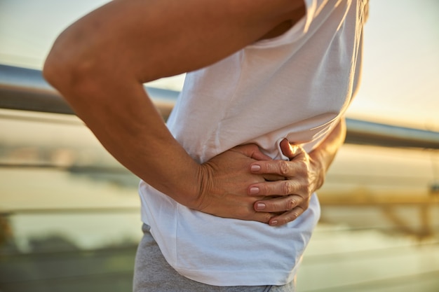 Close de uma senhora de camisa branca que sofre de dor abdominal no lado esquerdo enquanto passa um tempo ao ar livre