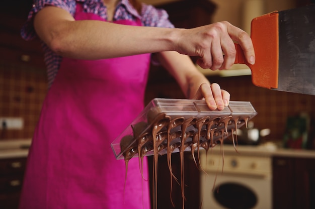Close do confeiteiro fazendo cascas para bombons de chocolate e removendo o excesso de chocolate dos moldes.