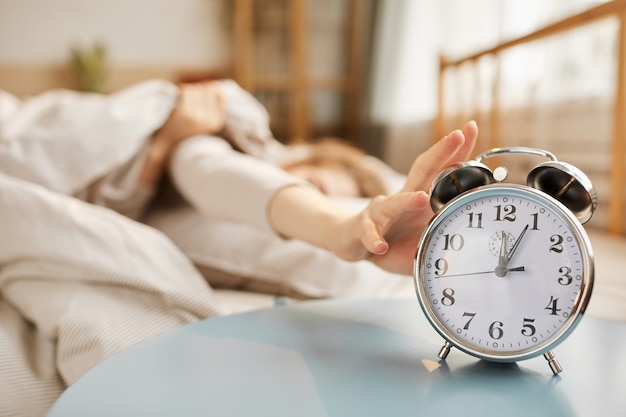 Close do despertador tocando na mesa com uma mulher dormindo na cama, na parede