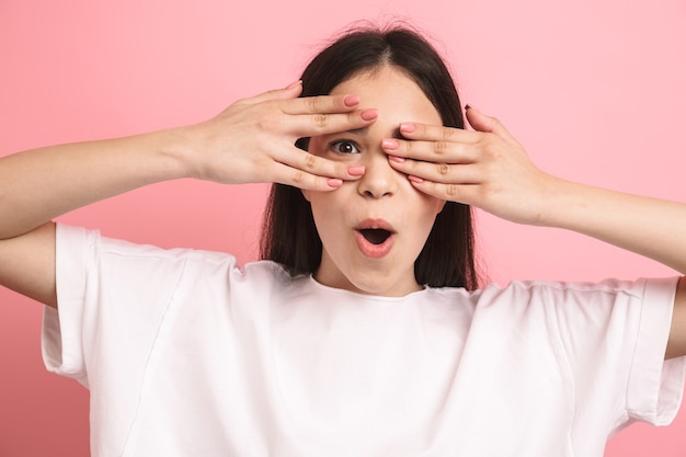 Close do retrato da doce menina positiva com longos cabelos escuros cobrindo os olhos e espiando por meio das mãos isoladas sobre a parede rosa