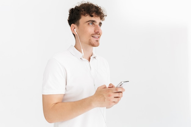 Close do retrato de um homem encaracolado caucasiano em uma camiseta casual sorrindo enquanto usa fones de ouvido e segurando o smartphone isolado sobre a parede branca