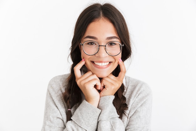 Close do retrato de uma adolescente alegre usando óculos, sorrindo e apontando os dedos para as bochechas isoladas sobre a parede branca