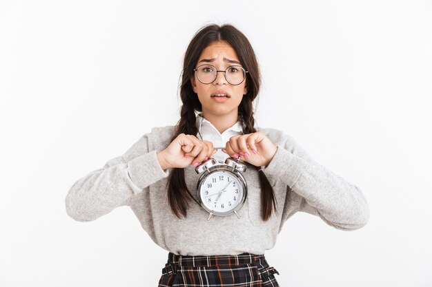 Close do retrato de uma adolescente perturbada usando óculos, franzindo a testa enquanto segura o despertador nas mãos, isolado sobre a parede branca