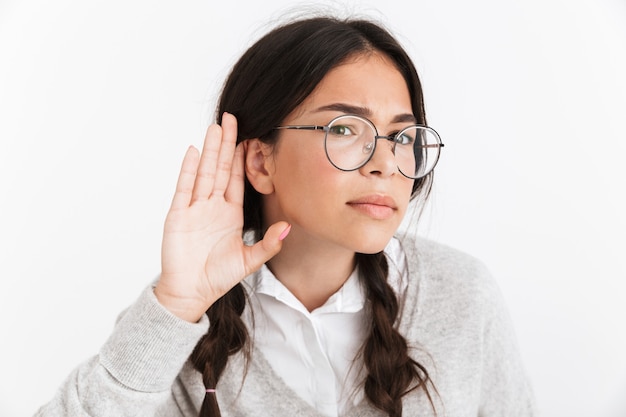Close do retrato de uma adolescente taciturna usando óculos, segurando a mão na orelha enquanto ouve ou escuta isolada na parede branca