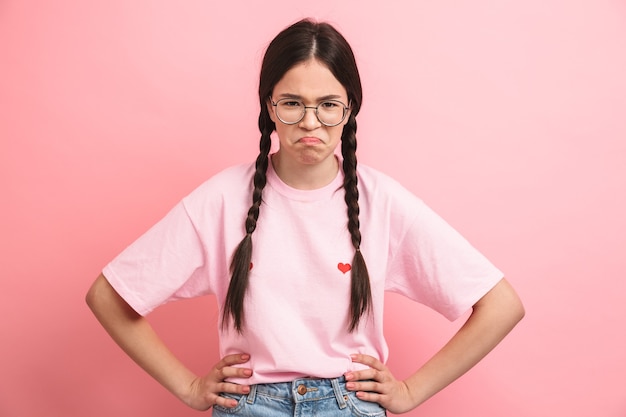 Close do retrato de uma divertida menina adolescente com duas tranças, usando óculos, se divertindo e fazendo careta na frente, isolada sobre a parede rosa