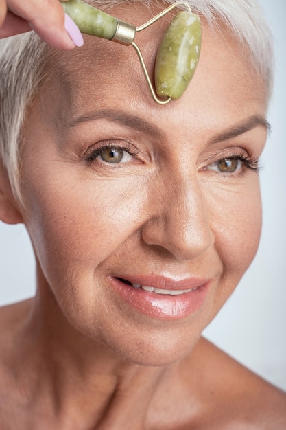 Foto close do rosto de uma mulher idosa rolando o rolo de jade na testa