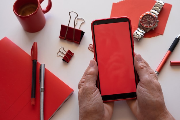 Close nas mãos de uma mulher usando telefone com tela vermelha. Área de trabalho branca e acessórios vermelhos