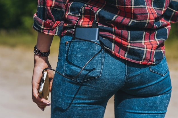 Close, Power Bank no bolso de trás da calça jeans da garota. Contra o pano de fundo da natureza.