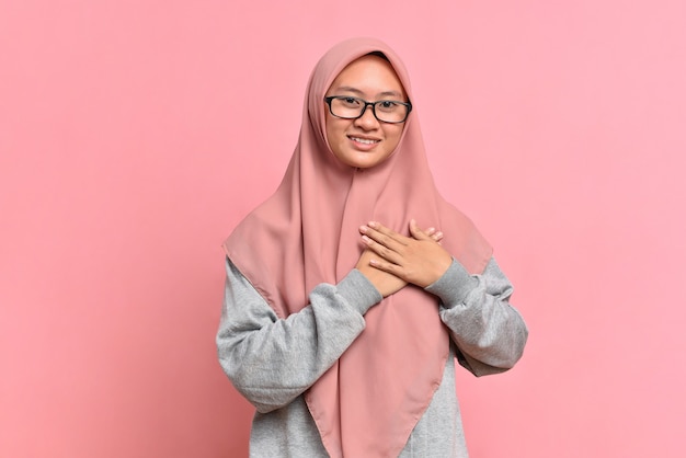 Close-up agradecido, mulher muçulmana asiática sentindo-se tocada e lisonjeada, sorrindo e olhando com afeto, tocar o coração, expressar gratidão e alegria por receber um belo presente, isolado no fundo rosa