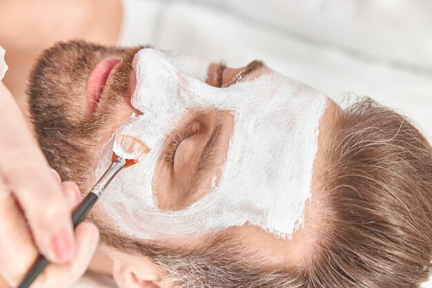 Close-up barbudo jovem está relaxando enquanto cosmetologista está espalhando argila branca no rosto.