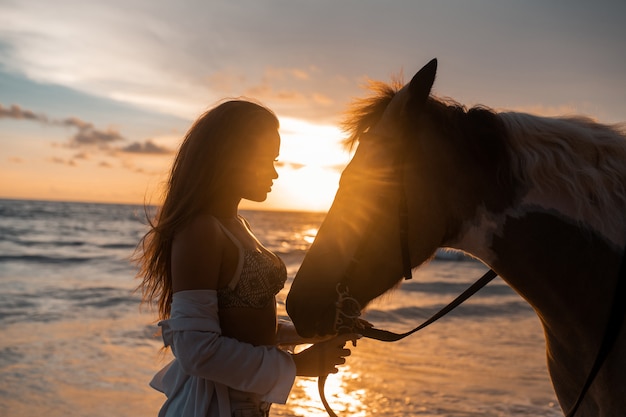 Close-up: beleza morena jovem se divertindo com o cavalo na praia. pôr do sol