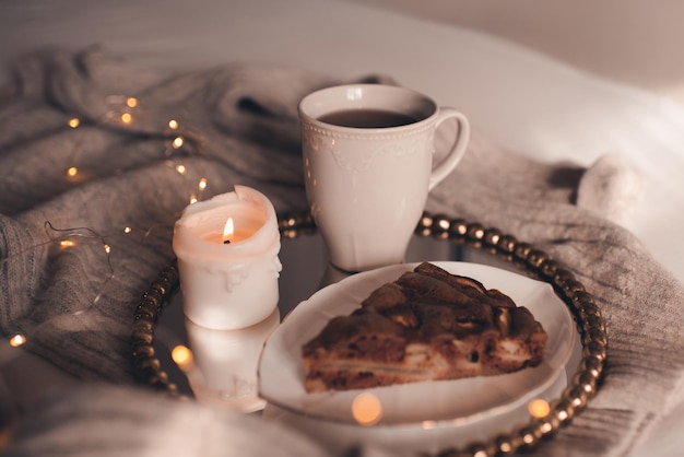 Close-up caneca de chá preto com vela acesa e torta de chocolate na cama sobre luzes de Natal. Bom Dia. Temporada de férias de inverno. Hora do lanche.