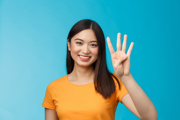Foto close-up carismático concurso elegante mulher asiática mostrar número quatro dedos, sorrindo feliz, fazendo o pedido, explicar quantos itens deseja comprar, ficar de pé fundo azul despreocupado