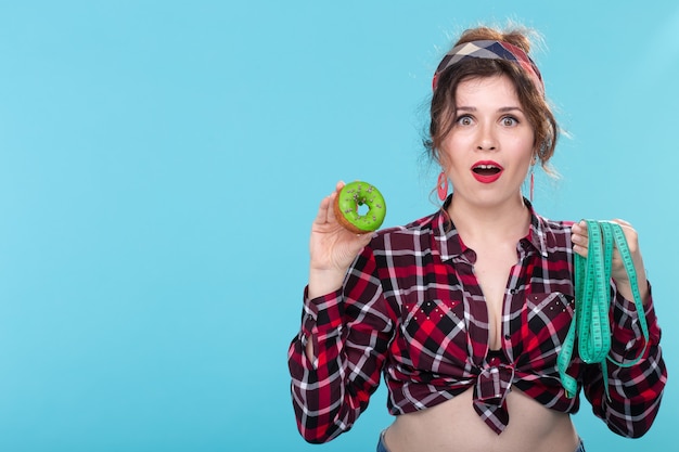 Close-up chocado surpreso jovem mulher bonita olhando para a fita métrica e segurando donut nas mãos, posando sobre fundo azul com espaço de cópia. Dieta e rejeição de alimentos nocivos com alto teor calórico.