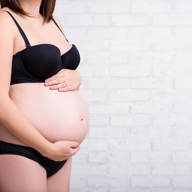 Close-up da barriga da mulher grávida em lingerie sobre a parede de tijolos brancos