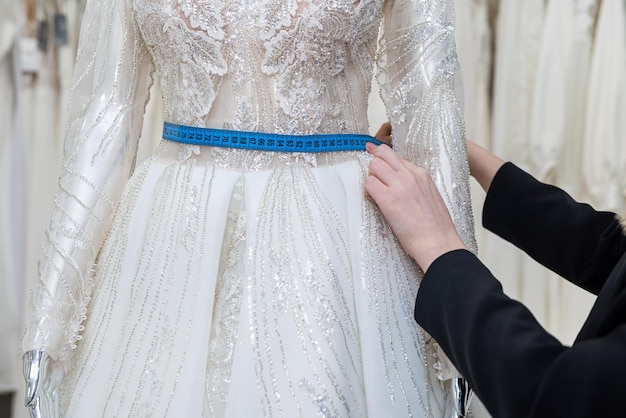 Close-up da costureira de estilista feminina que ajusta o vestido de noiva com fita métrica na boutique