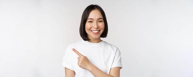 Close-up da jovem japonesa sorrindo apontando o dedo para a esquerda no espaço da cópia mostrando anúncio ou banner de propaganda em pé sobre fundo branco