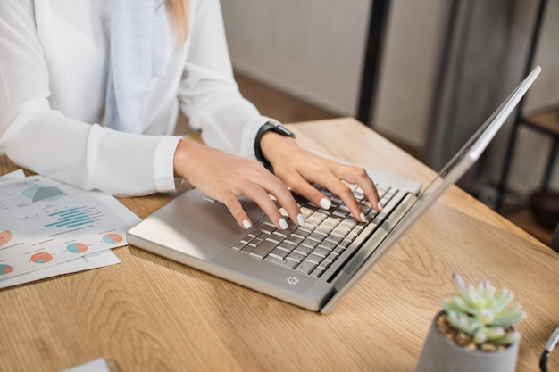 Close-up da mão da mulher com roupa formal com relógio inteligente no pulso digitando no laptop moderno